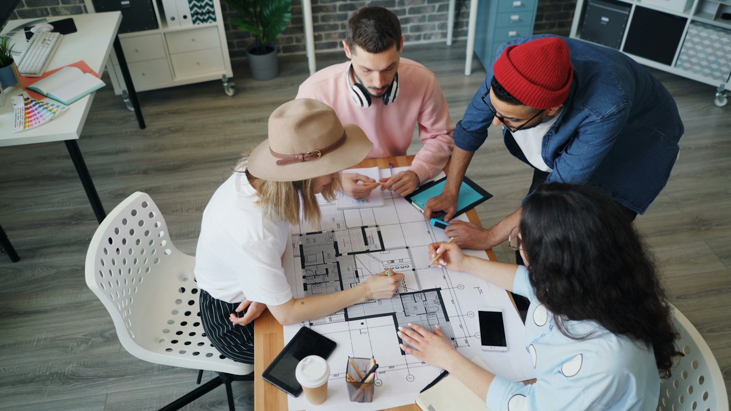 Fire voksne diskuterer tegninger ved et trebord.